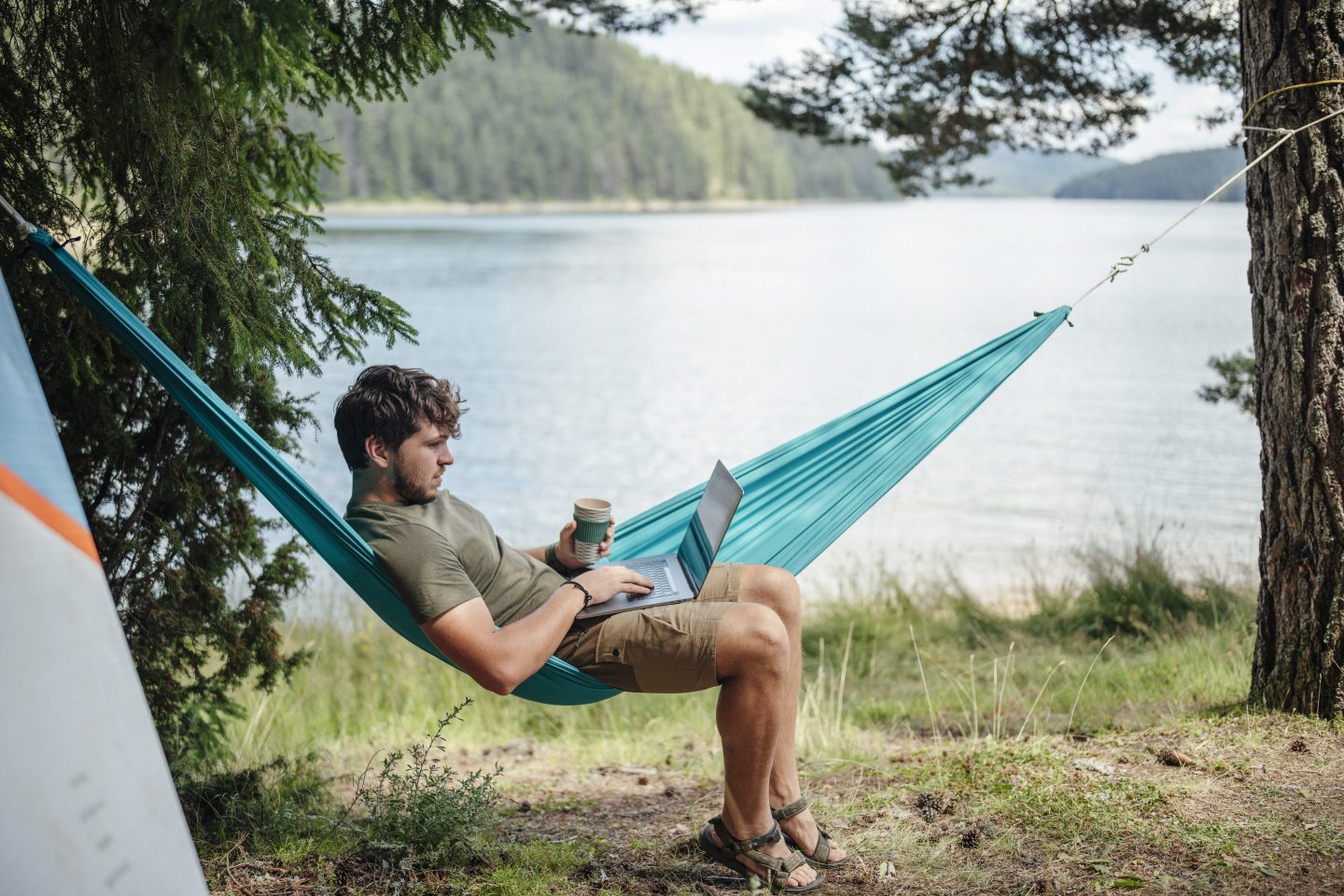 A man works on a laptop while lounging in a hammock in front of a lake.