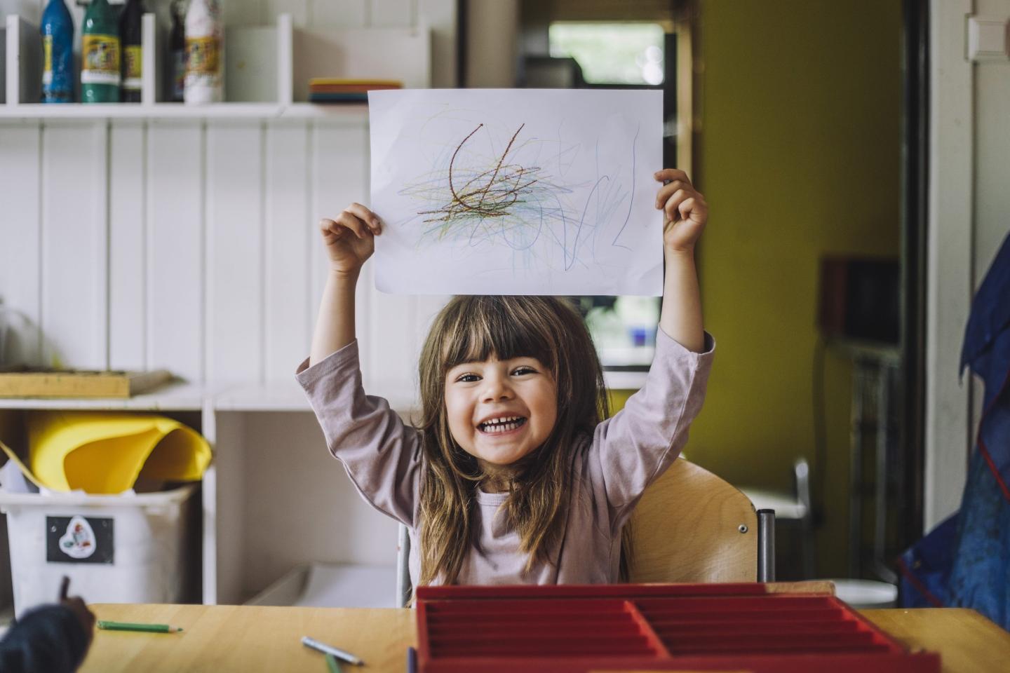 Portrait of happy girl showing color pencil scribble on paper at kindergarten