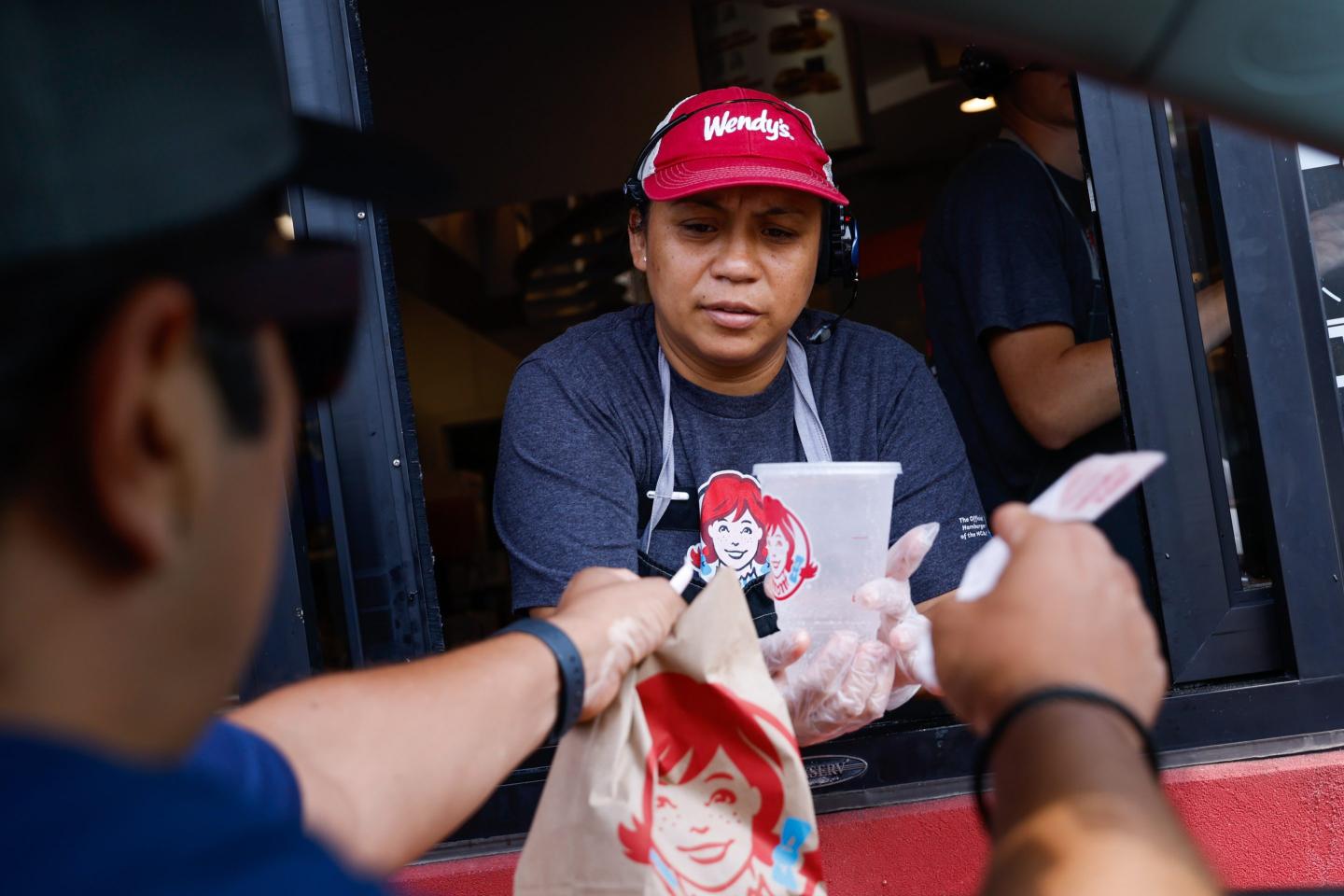 A Wendy's drive-thru worker hands a brown paper bag and drink to a customer in a car.
