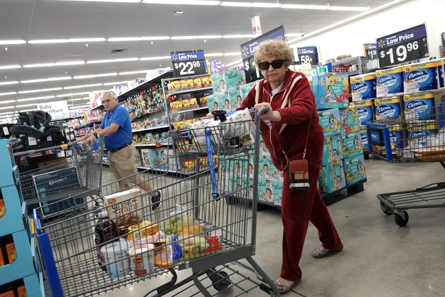 An older Walmart shopper in a red jumpsuit and sunglasses pushes a shopping cart down an aisle.