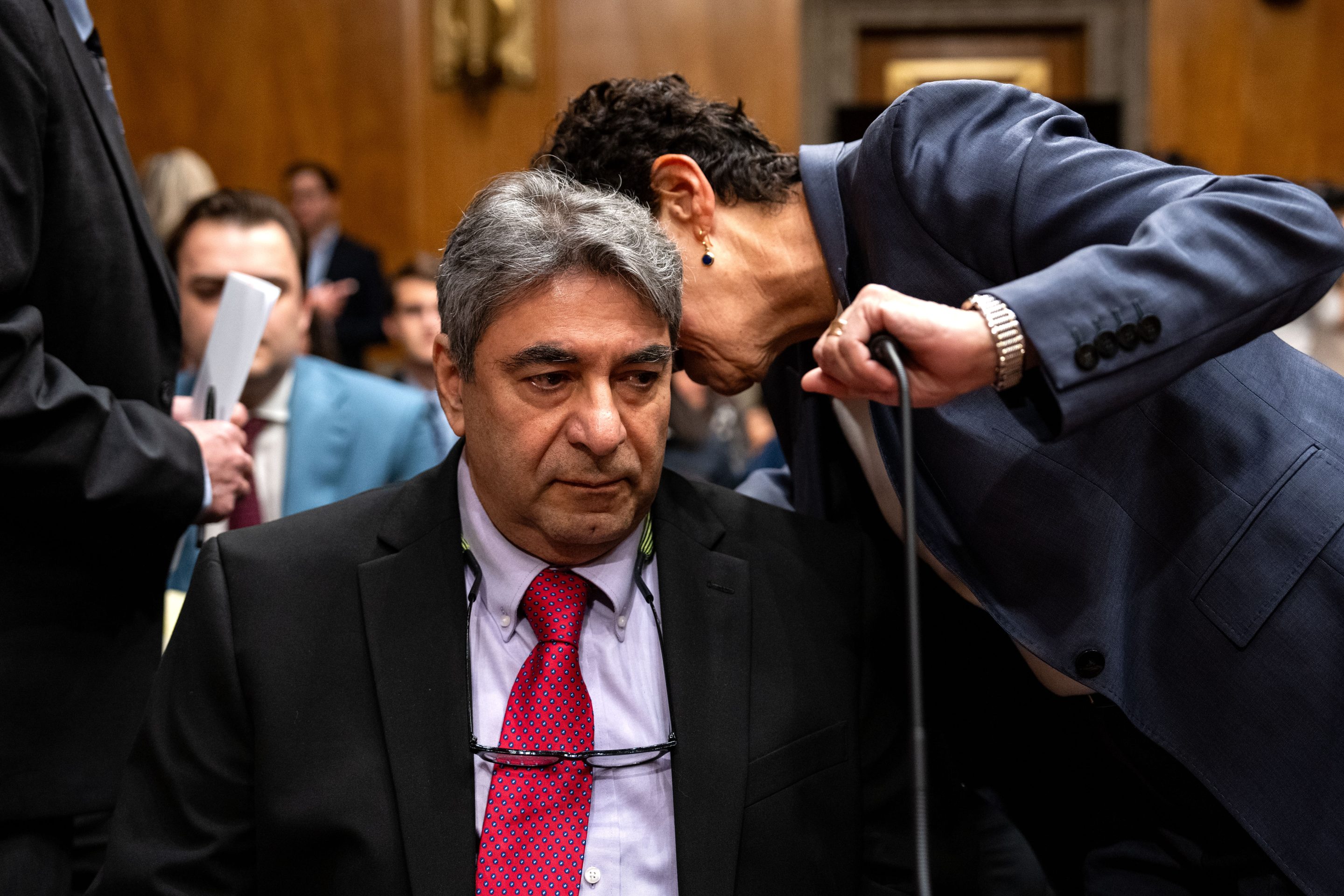 A woman is leaning over to whisper something to Sam Salehpour and is covering a microphone in front of him with her hand.