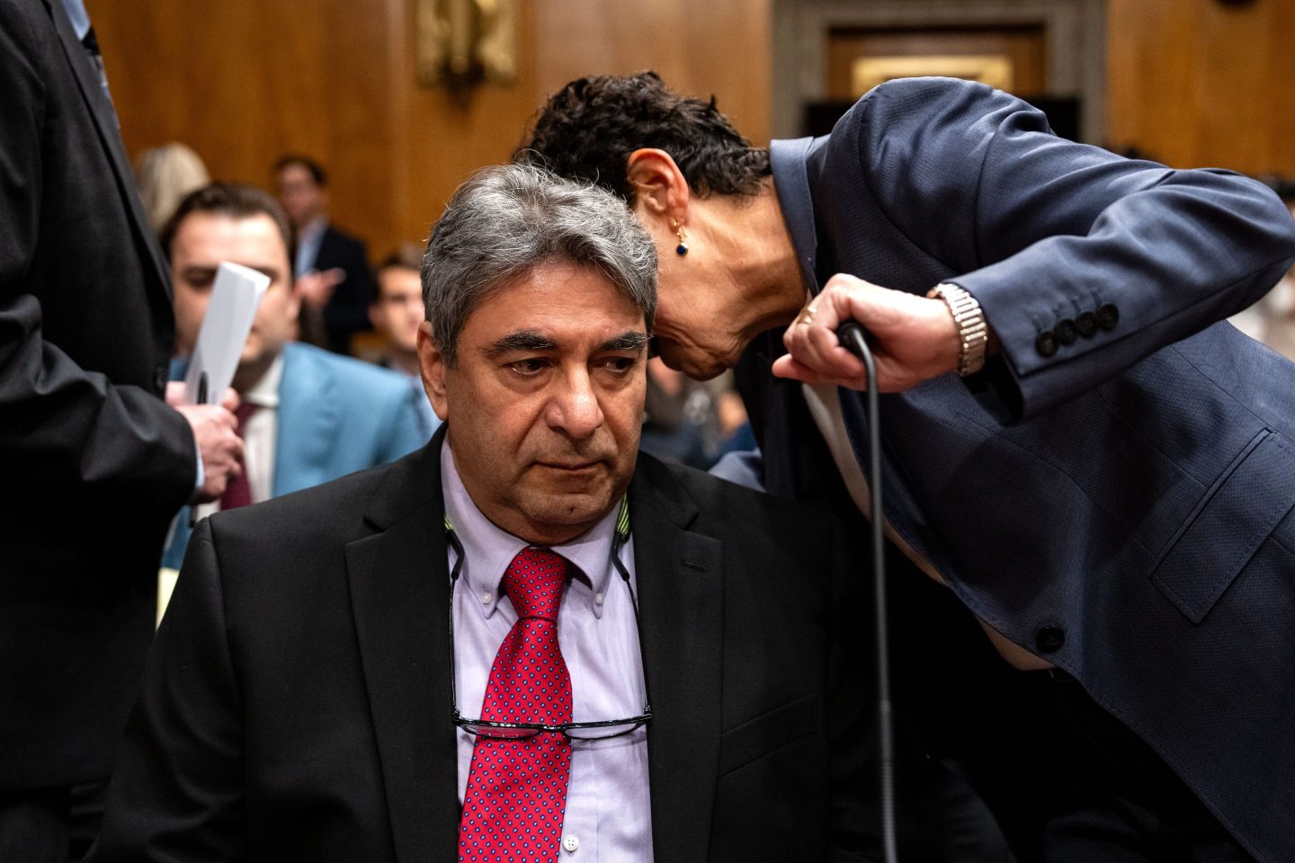 A woman is leaning over to whisper something to Sam Salehpour and is covering a microphone in front of him with her hand.