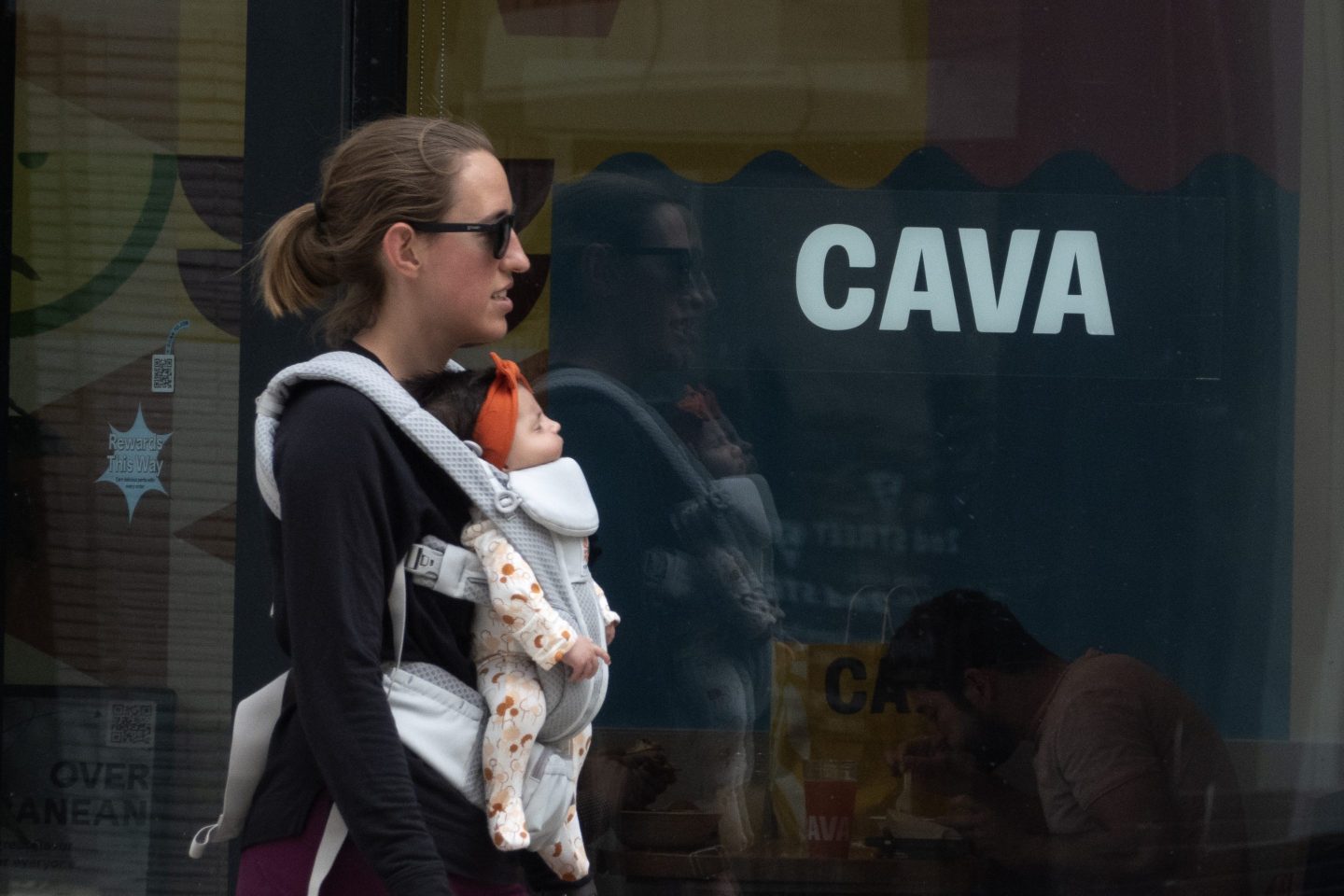 A women with a baby strapped to her chest walks past a window with the "Cava" logo across it.