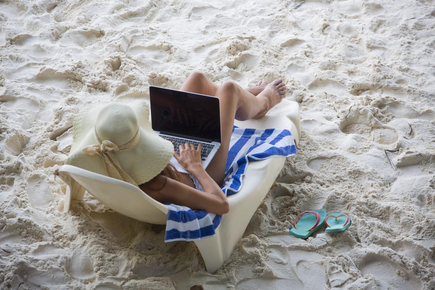 A woman sits on a lounge chair on the beach while working on her laptop.