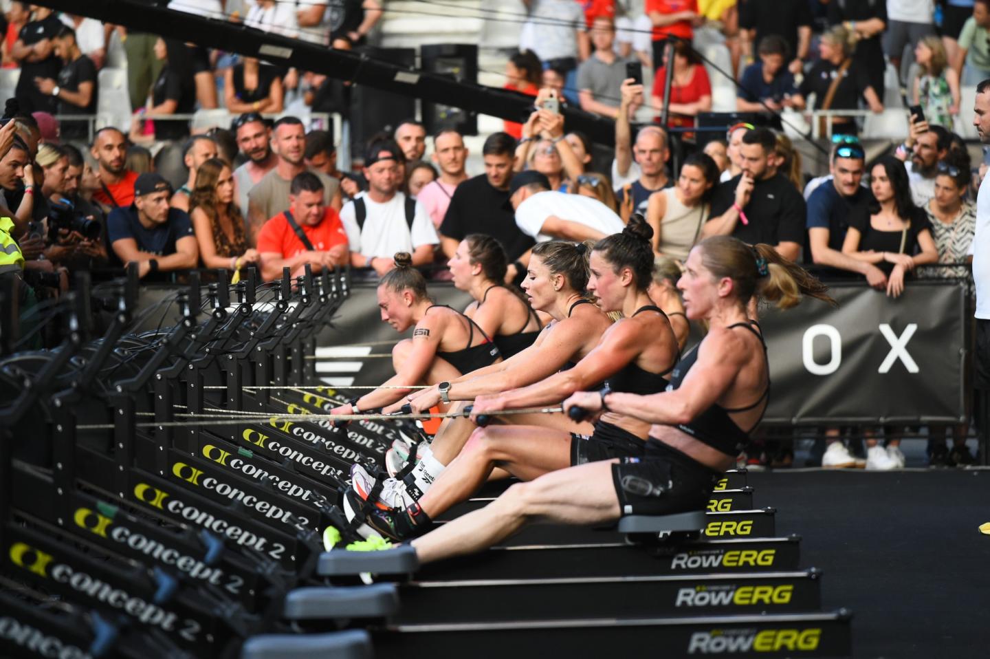 a row of women on the rowing machine with people watching at the background