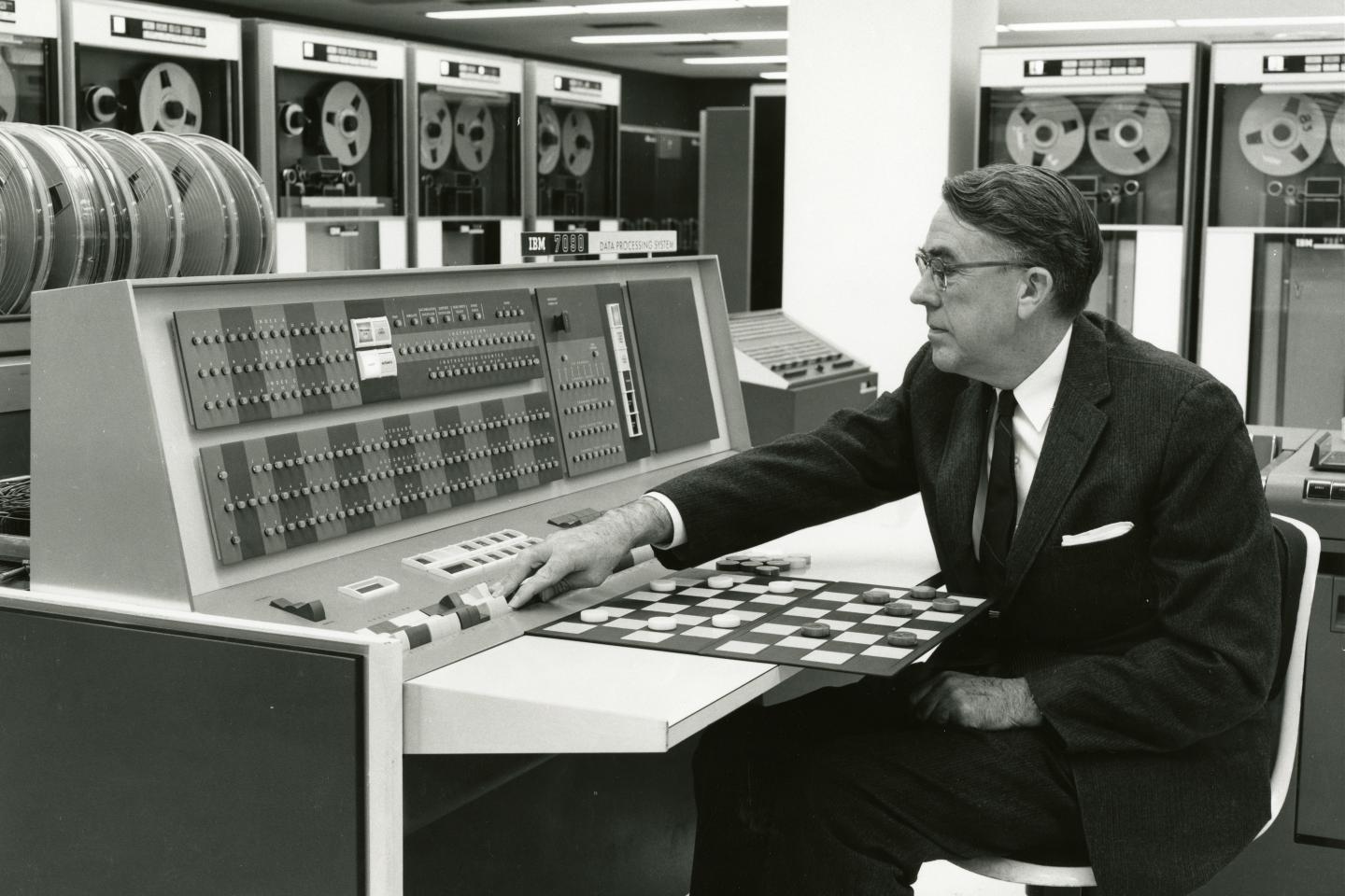 A middle-aged man in a suit sits in front of a checker board, working a computer's control panel.