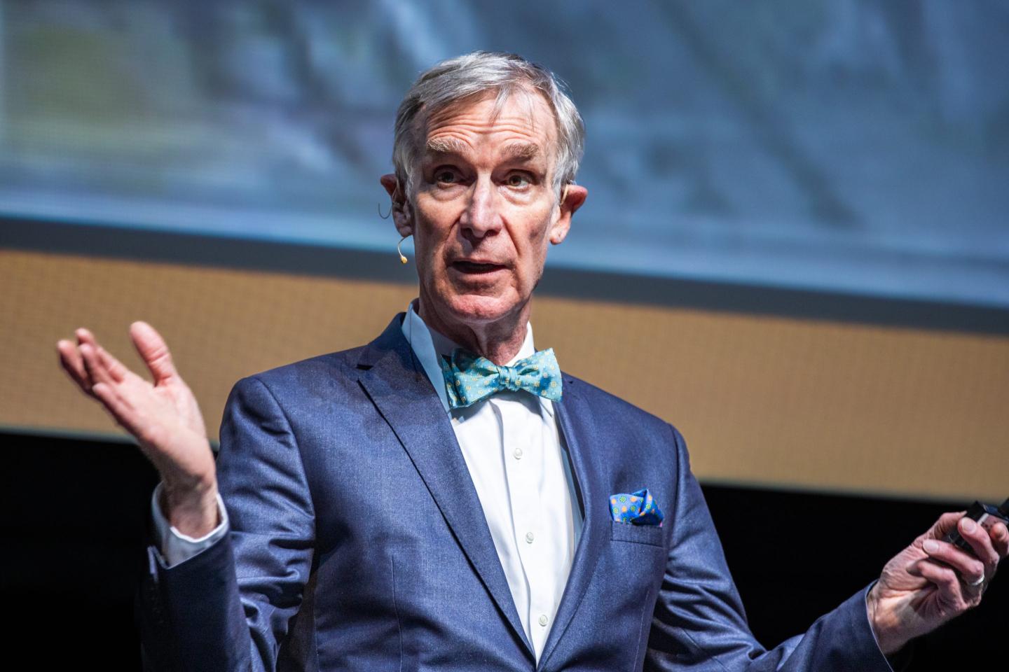 Bill Nye the Science guy speaks at an event, wearing a teal bowtie and blue suit.