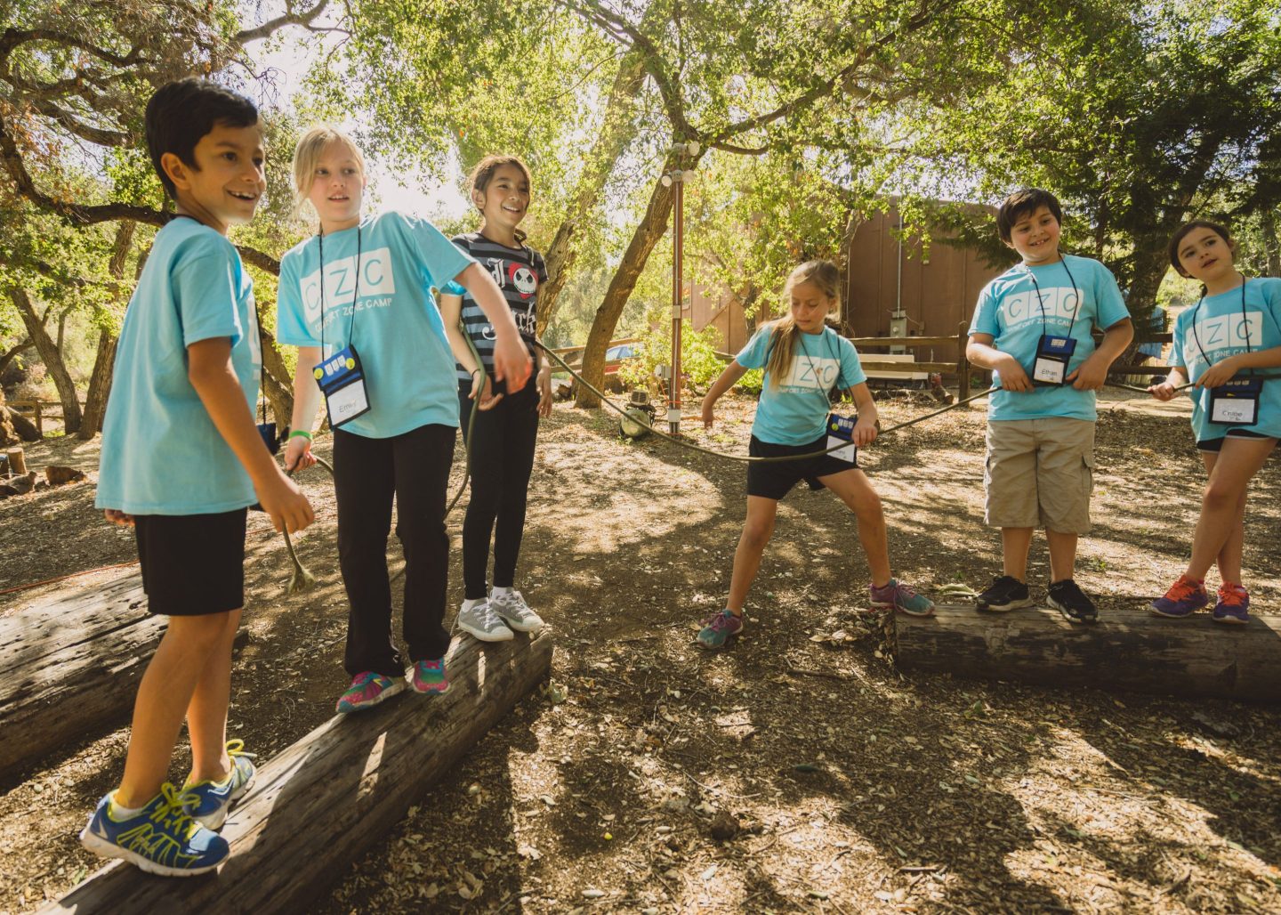 Six young campers in bright camp t-shirts stand on logs in the woods.