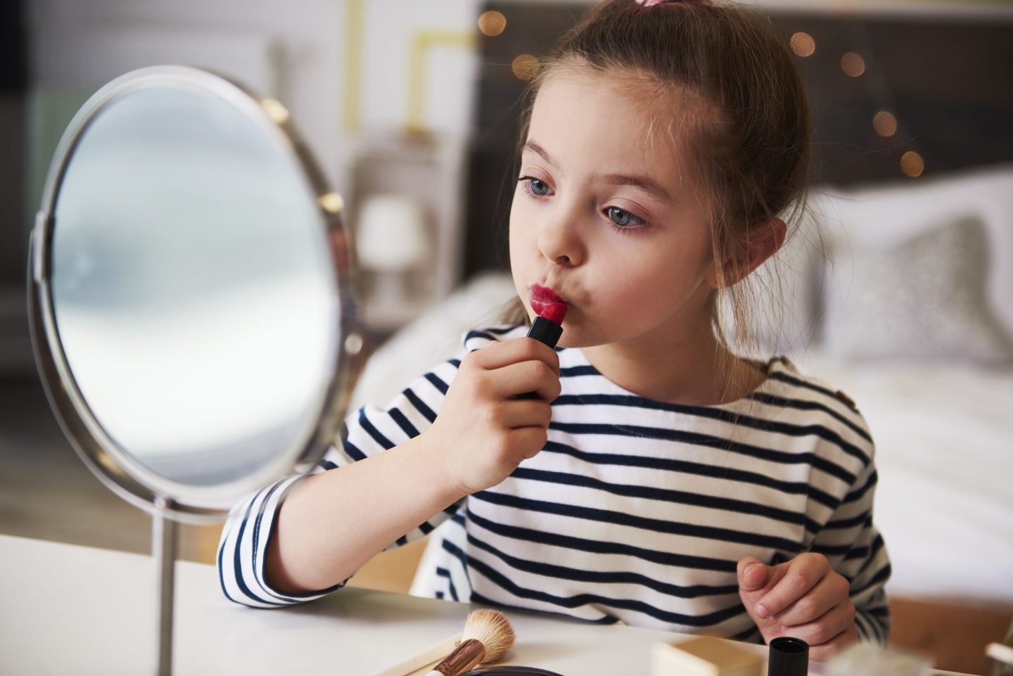 A young girl applies red lipstick in front of a mirror