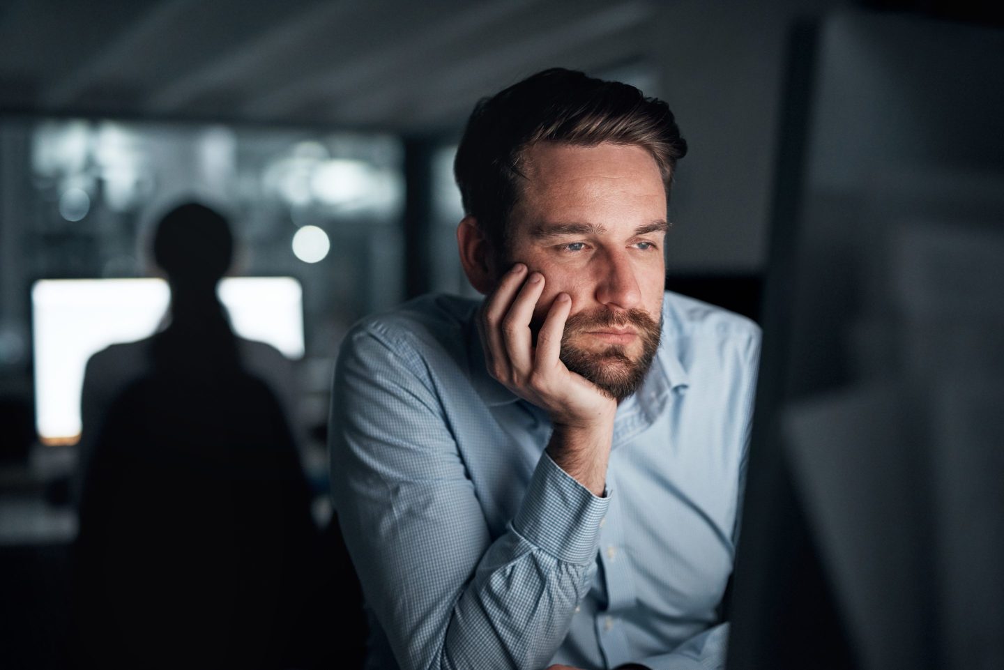 A man sits in a dark office staring at a brightly-lit computer screen. He looks bored.