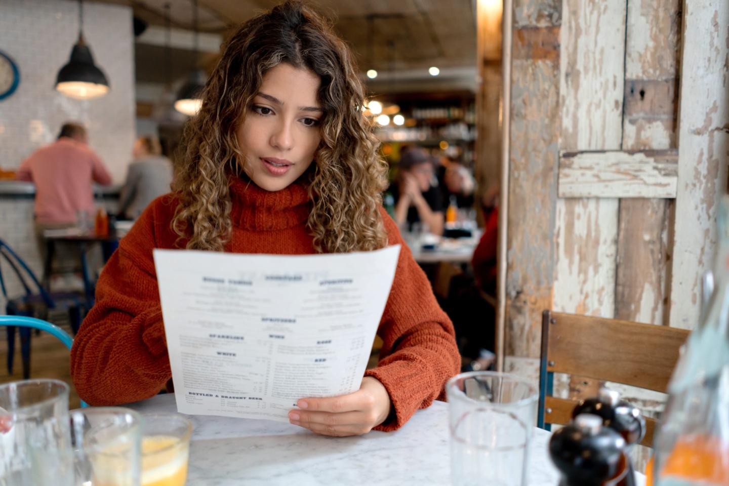 A woman is sitting in a cafe looking down at a resume she is holding.
