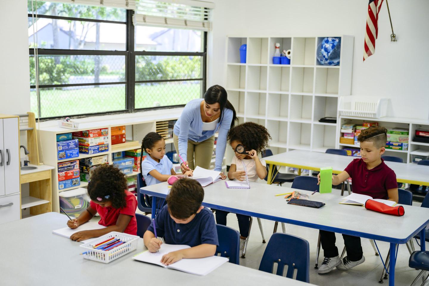 A young woman teacher is bending over at a table at school where young students are working