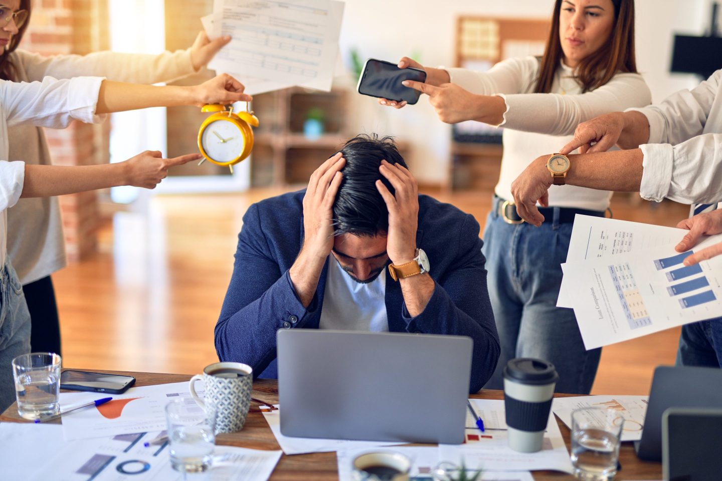 A man dressed in business casual attire sits at a messy desk and looks distraught, with his head in his hands. There is a group of workers surrounding him holding up various papers or items, trying to get his attention.