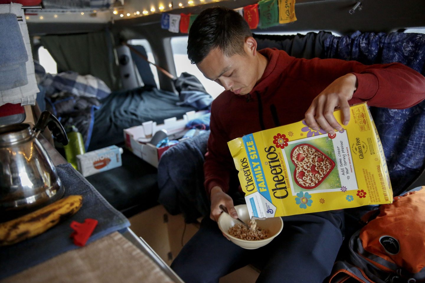 A man sits in a van pouring Cheerios from a box into a bowl.
