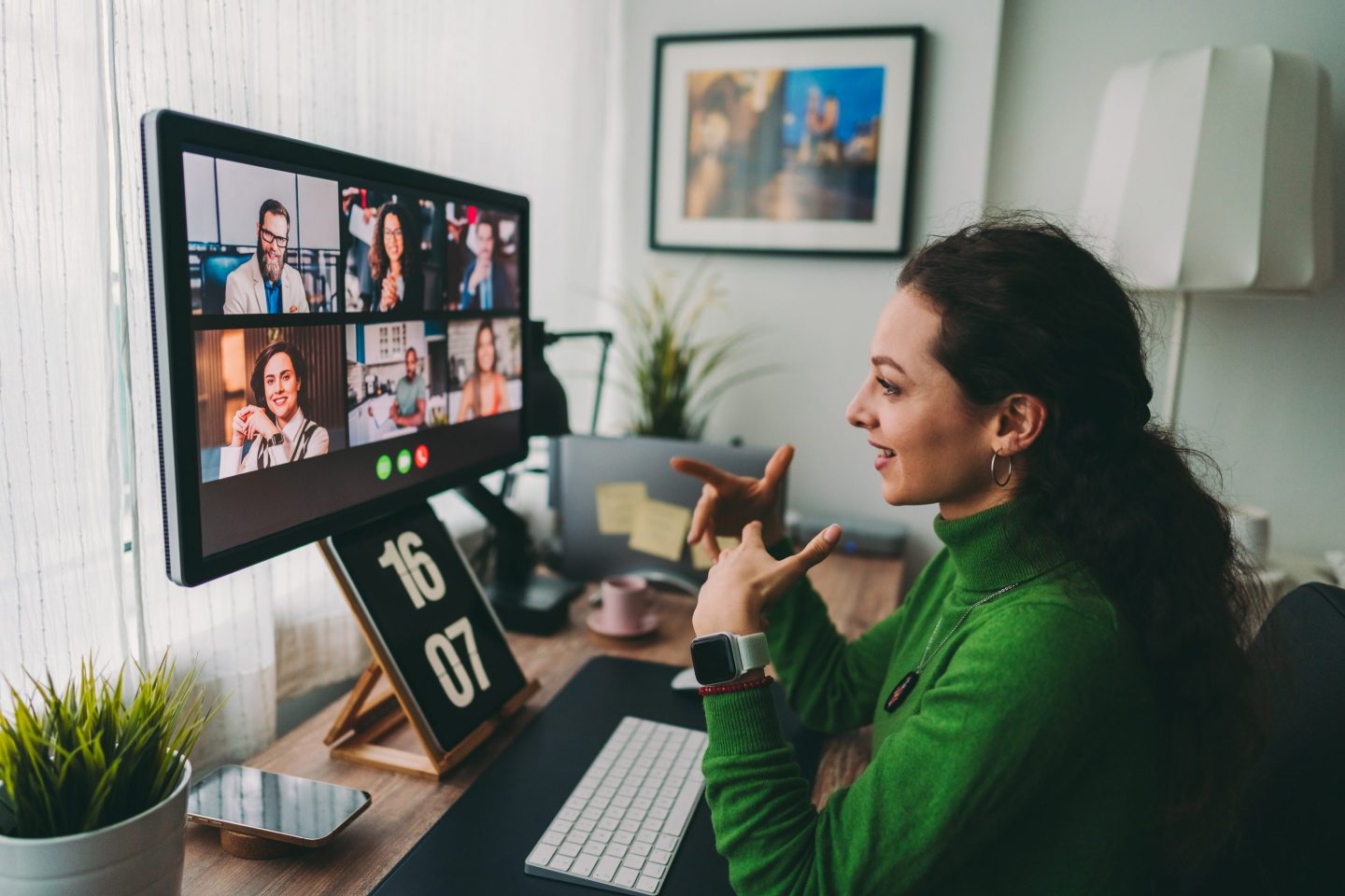 A woman in a green turtleneck sits at a desk on a telecall.
