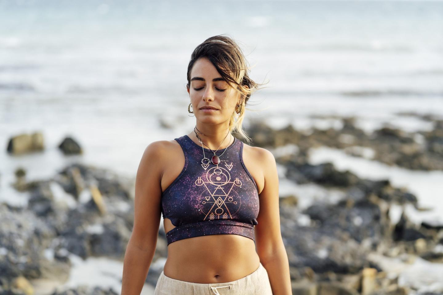 A women wearing a zodiac crop top stands on the beach with her eyes closed.