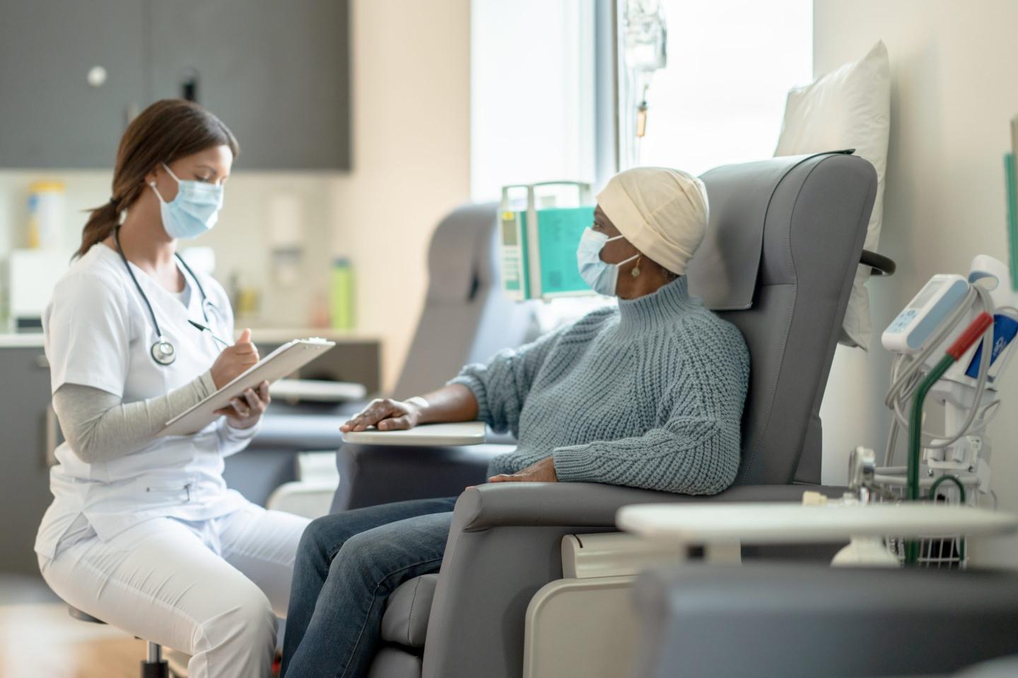 A woman in a knit cap sits in a chemo chair while a doctor crouches at her side