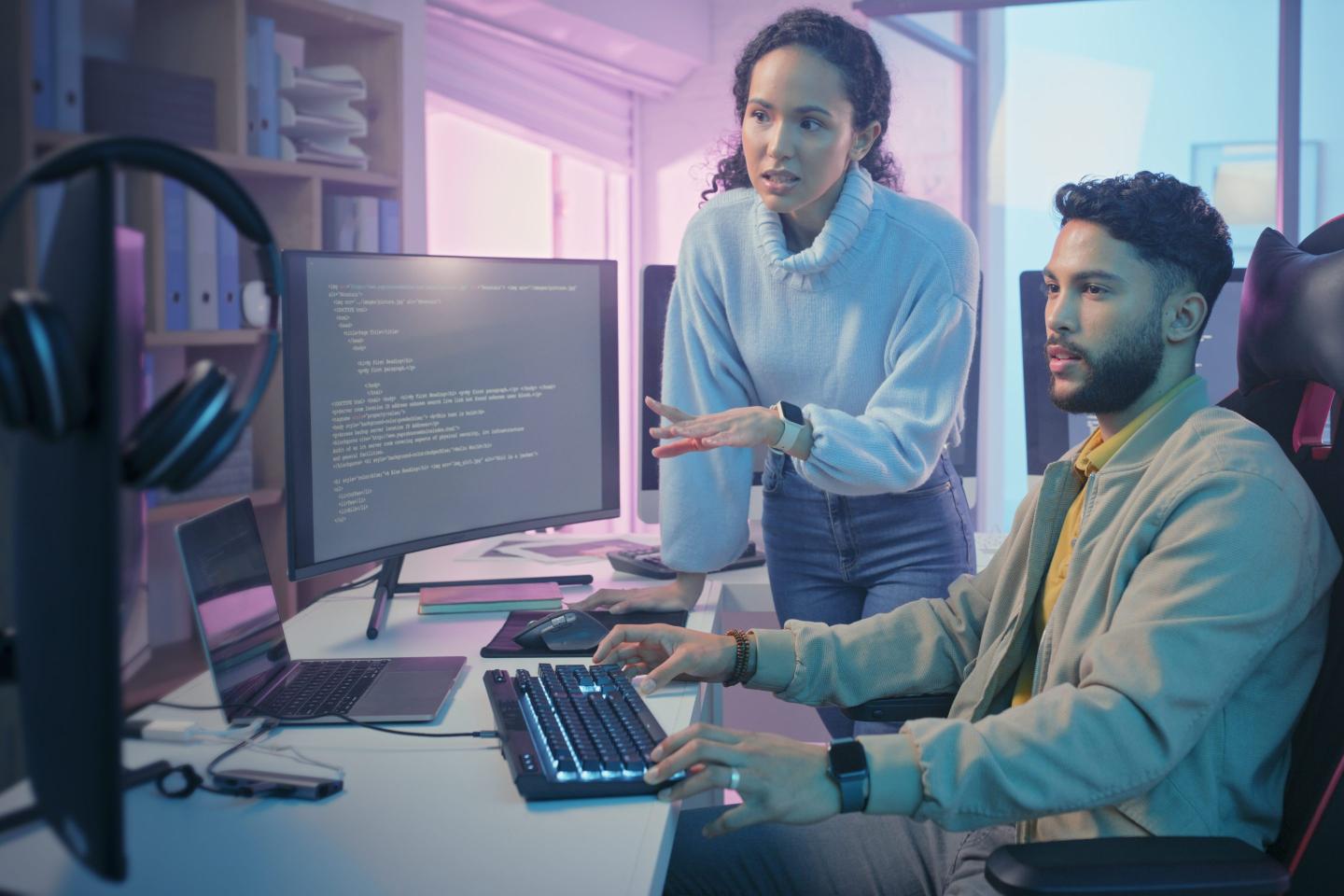 A woman in business casual attire stands next to a man in business casual attire sitting at his desk. They are looking at his computer screen.