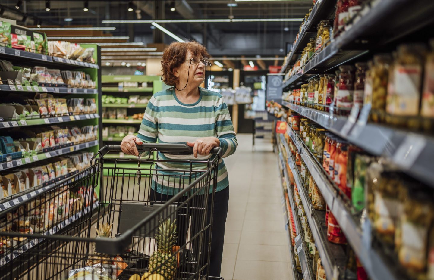 An older woman with a shopping cart walks down a grocery store aisle.