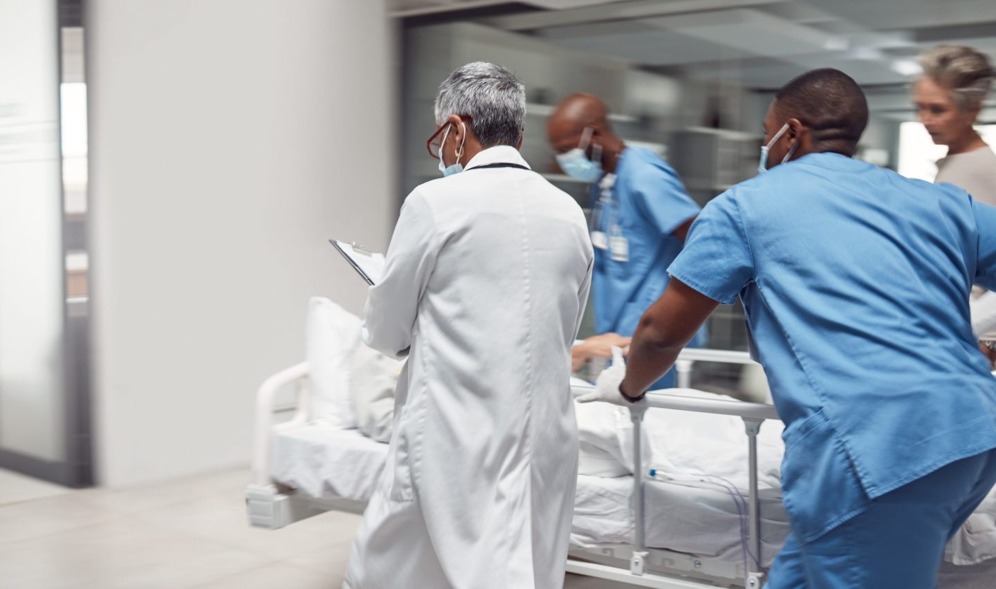 Image of a doctor in a white coat and several people in blue scrubs pushing a patient on a gurney in an emergency room.