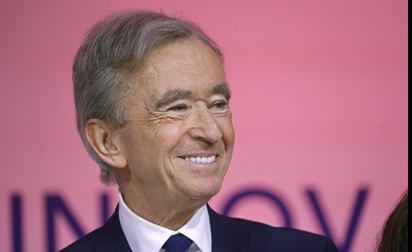 Bernard Arnault smiles to his right and is in front of a pink background.