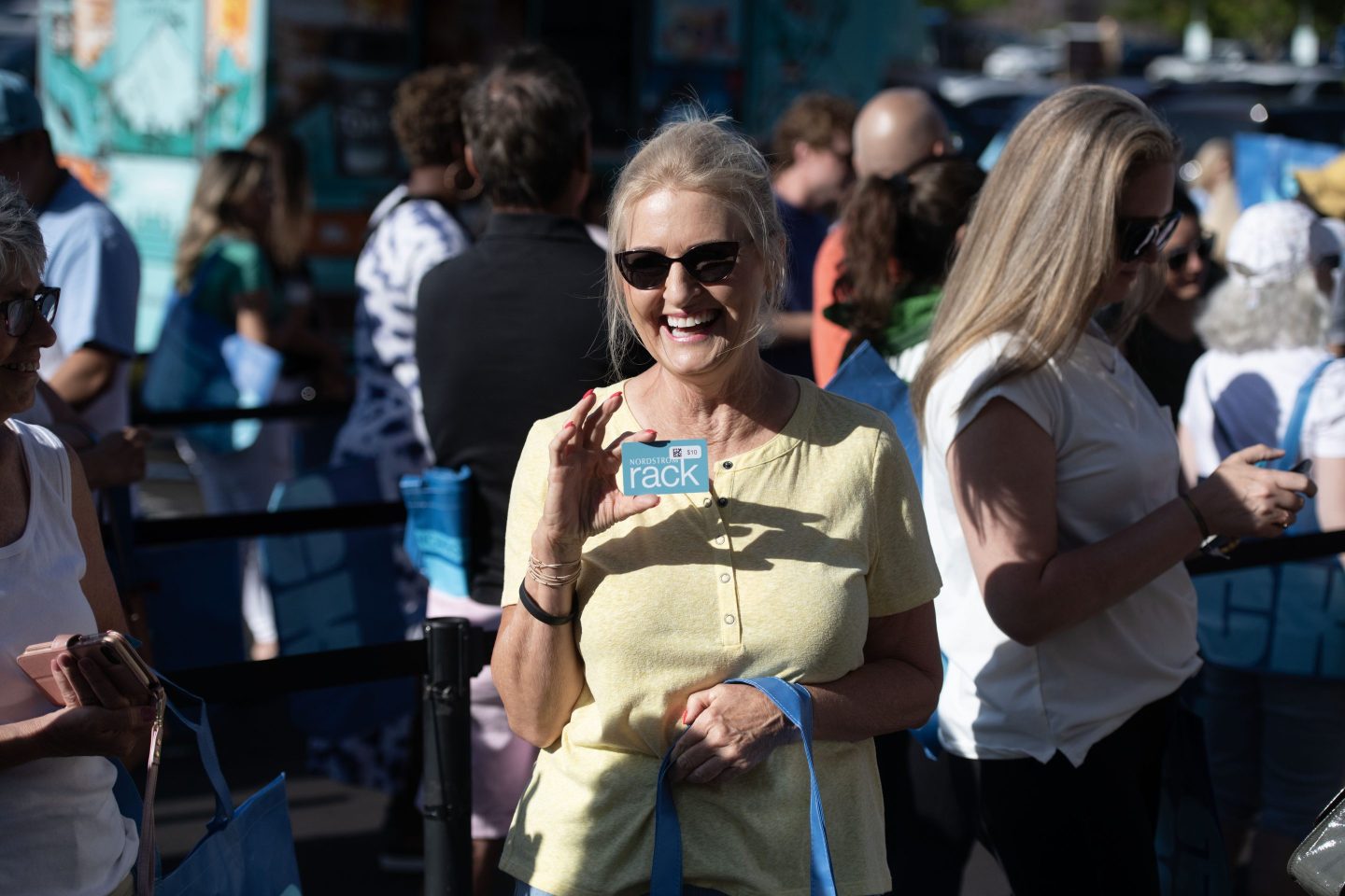 A woman stands in a crowd and holds up a Nordstrom Rack credit card to the camera, smiling.