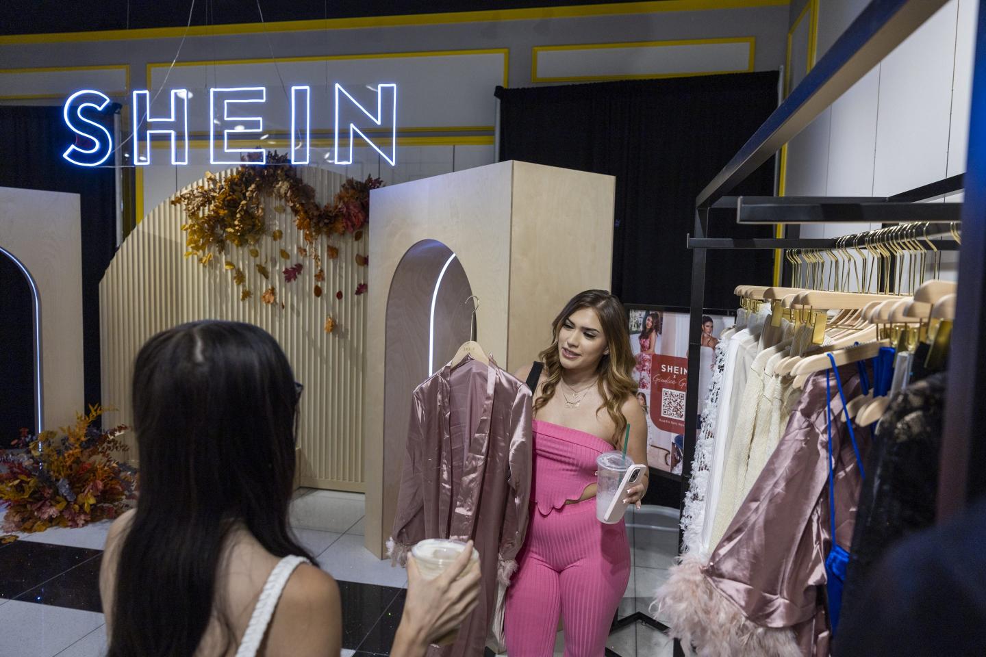 A woman in a pink jumpsuit holds up a pink garment to show her friend