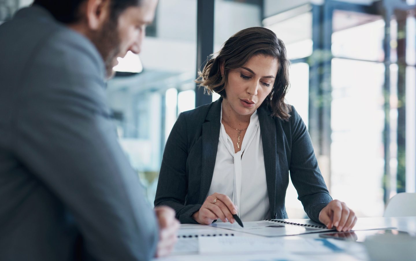 A businessman and woman sit at a meeting table in an office, reviewing and discussing documents in front of them.