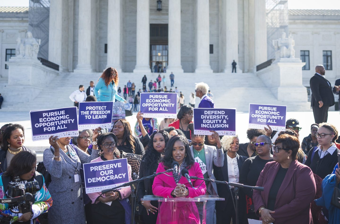 Arian Simone, in a pink suit, addresses a crown in front of the Supreme Court