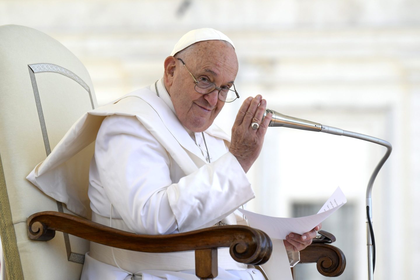 Pope Francis, dressed in all white, sits in a chair and covers a microphone with his hand.