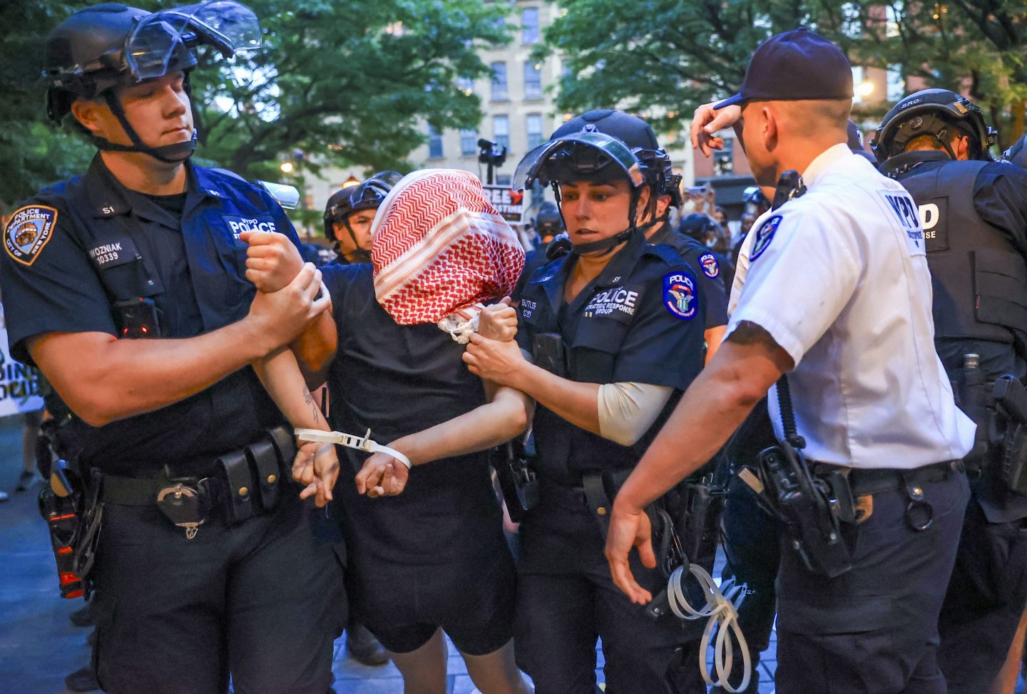Two NYPD police officers detain a pro-Palestinian activist whose face is hidden by a keffiyeh.