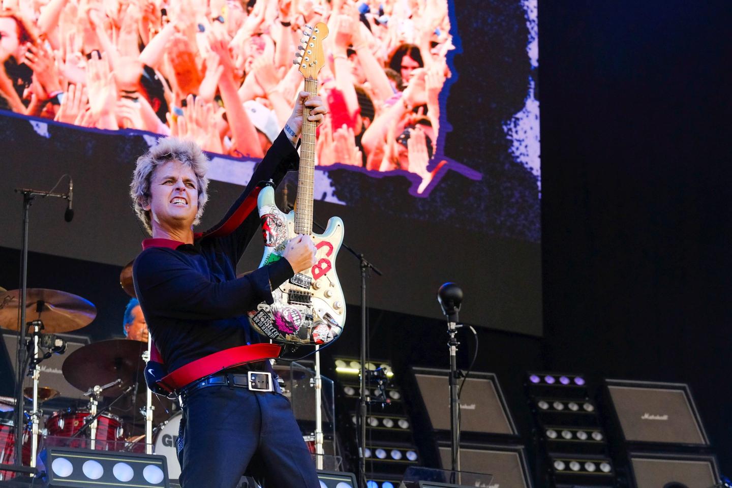 Guitarist and vocalist Billie Joe Armstrong brandishing a guitar on stage in front of a screen showing images of a large audience as he performs live with American punk band Green Day at the Isle of Wight Festival.