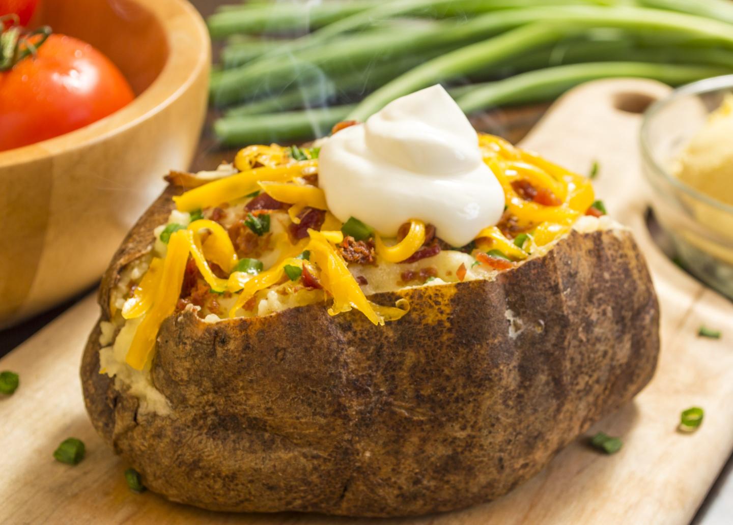 fresh hot baked potato with butter bacon cheese and chives topped with sour cream on a cutting board