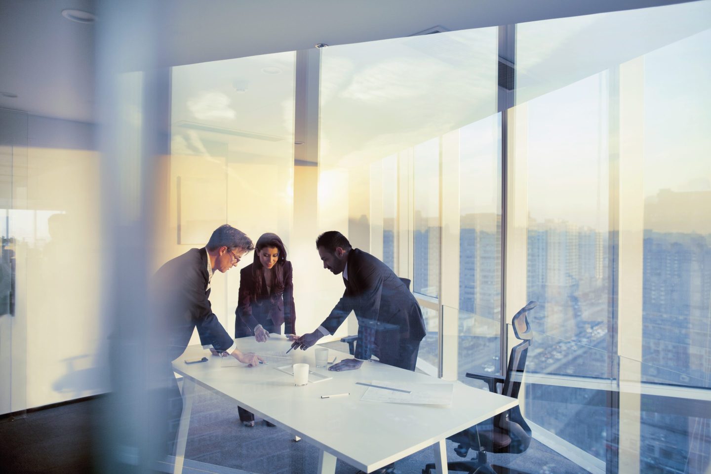 A group of business people stand around a meeting table discussing documents in front of them.