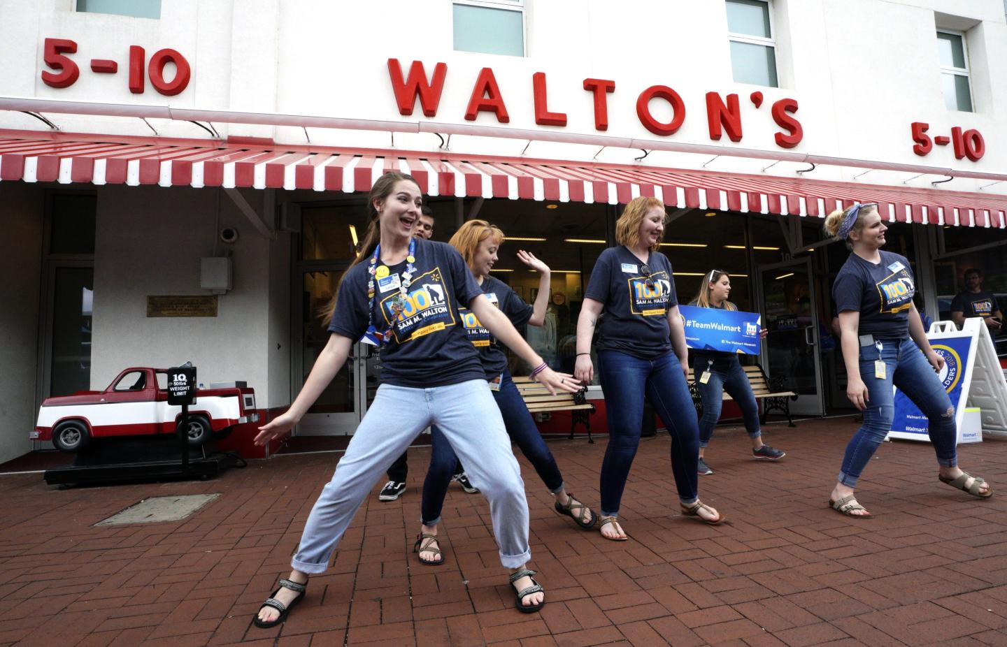 A group of young women dance in front of a white-and-red storefront with a "Walton's" sign across it.