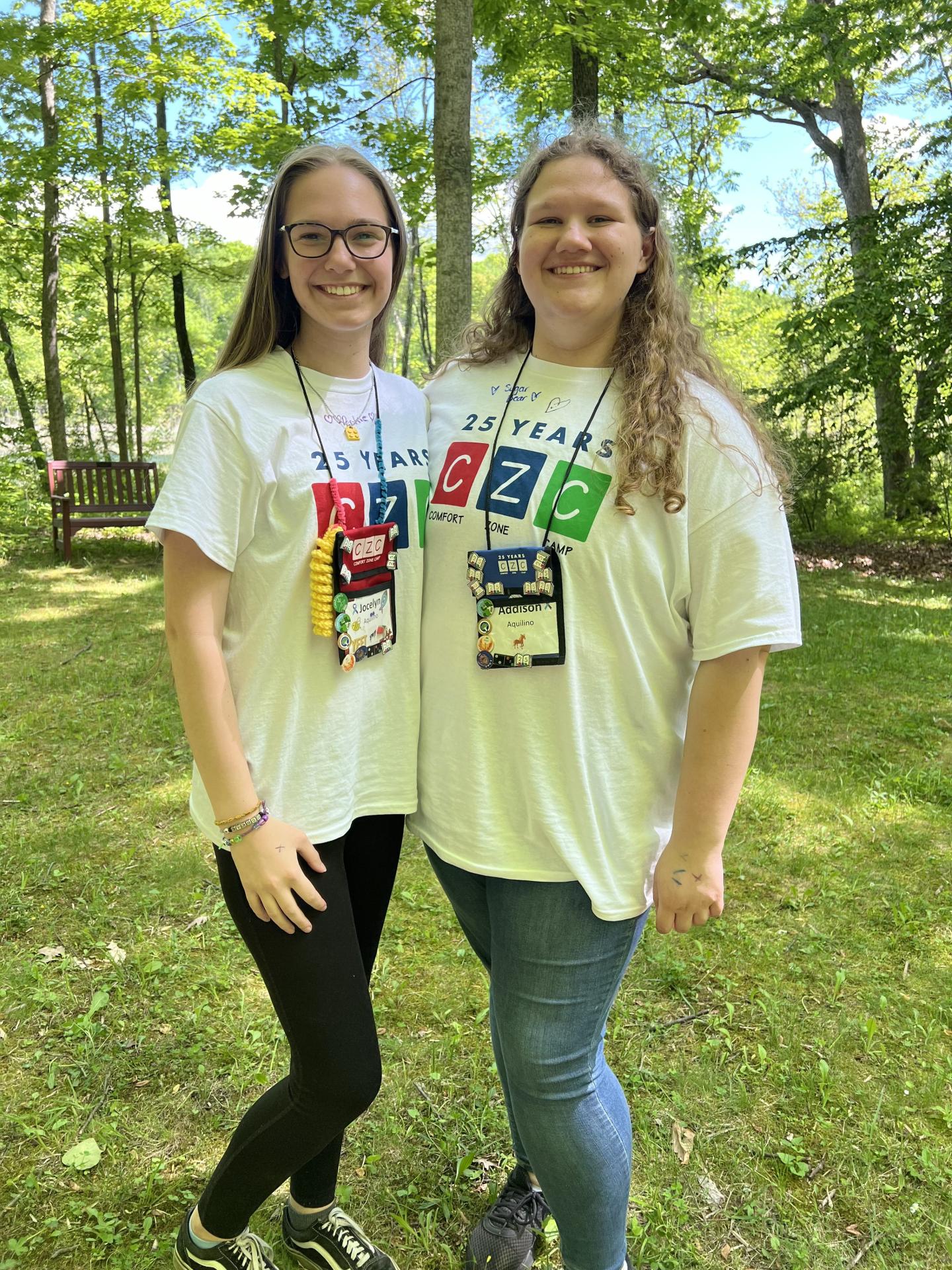Two sisters smile and pose next to each other in the woods wearing white camp t-shirts and lanyards.