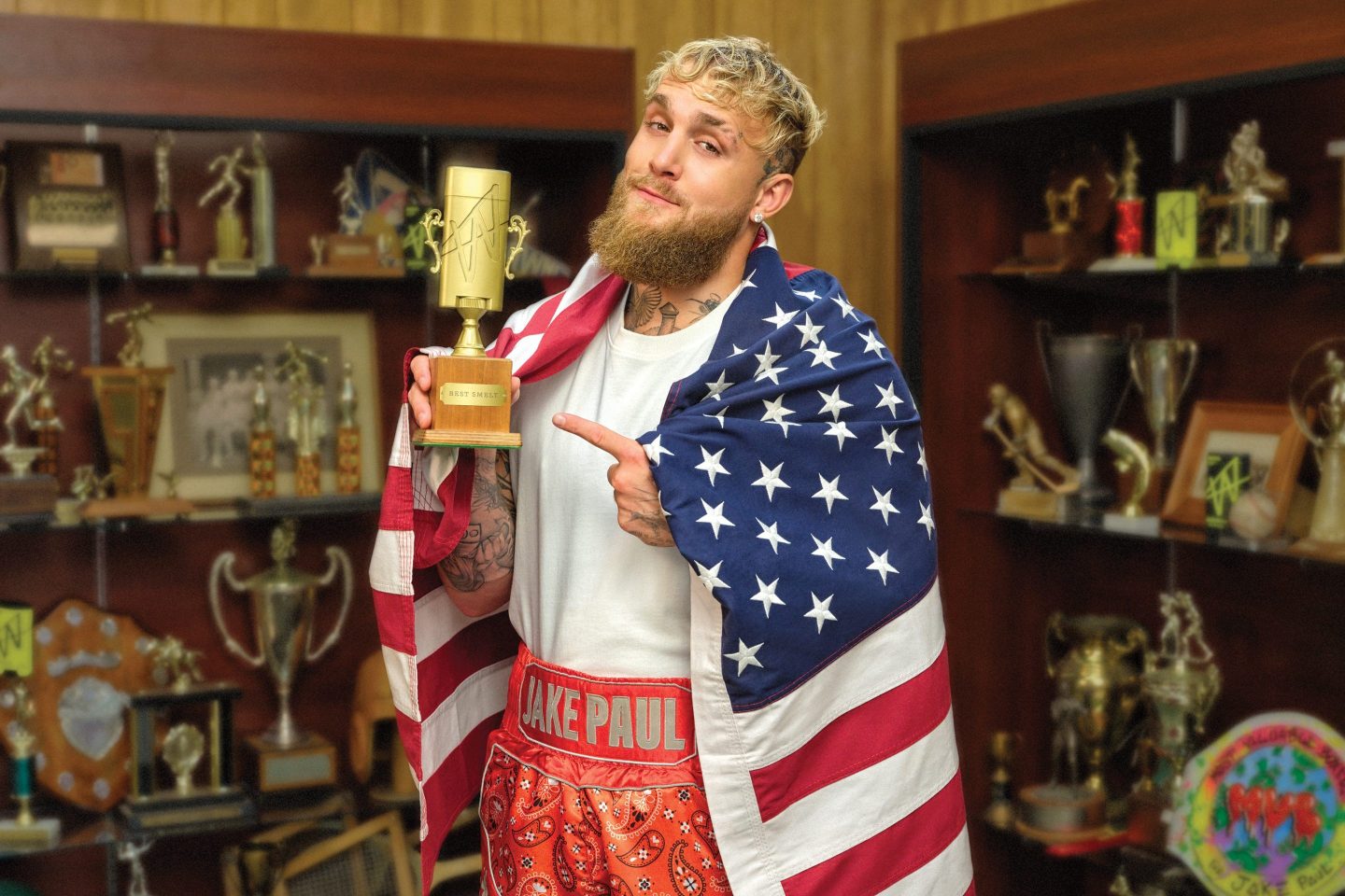 Jake Paul is standing in room lined with trophies. He is wrapped in an American flag and holding a gold trophy in the shape of a deodorant tube, pointing at it.