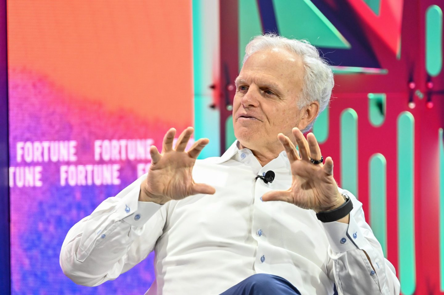David Neeleman speaks with his hands in front of him while sitting in front of a colorful backdrop.