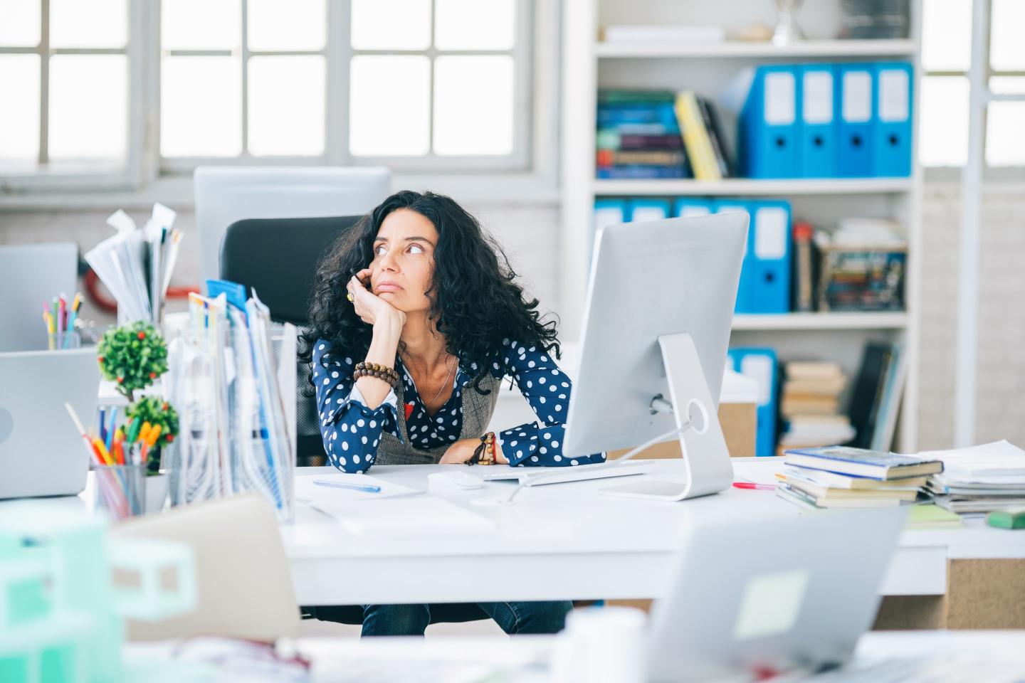 A woman sits at a cluttered desk with her hand in her palm as she looks to the side.
