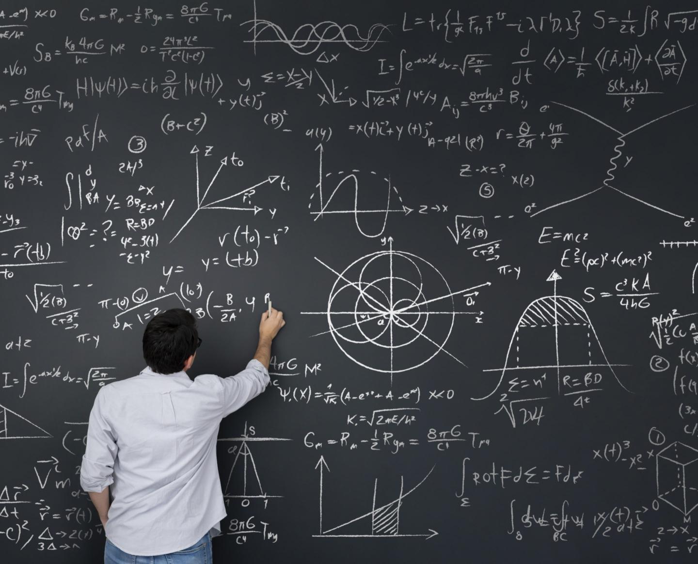 A man standing at a blackboard filled with equations and diagrams writing with chalk on the board.