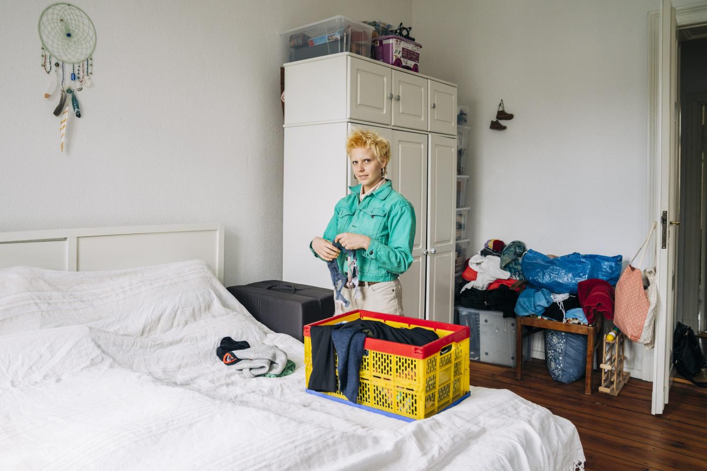A woman stands next to her bed while folding clothes into a crate.