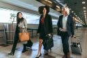 Three business people walk together in an airport terminal, carrying luggage.