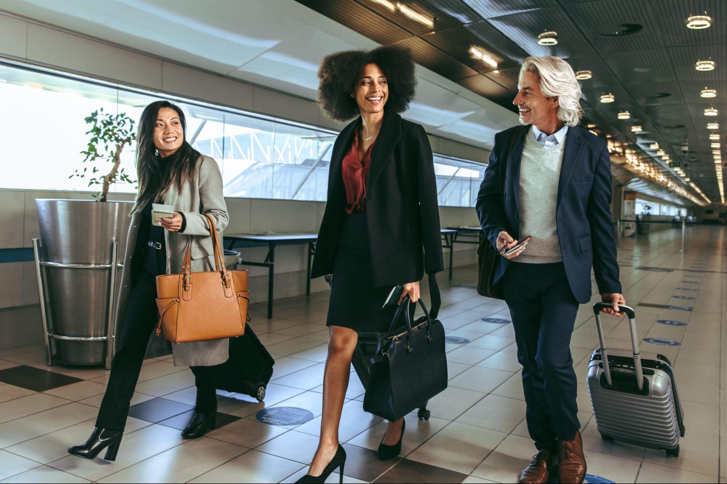 Three business people walk together in an airport terminal, carrying luggage.
