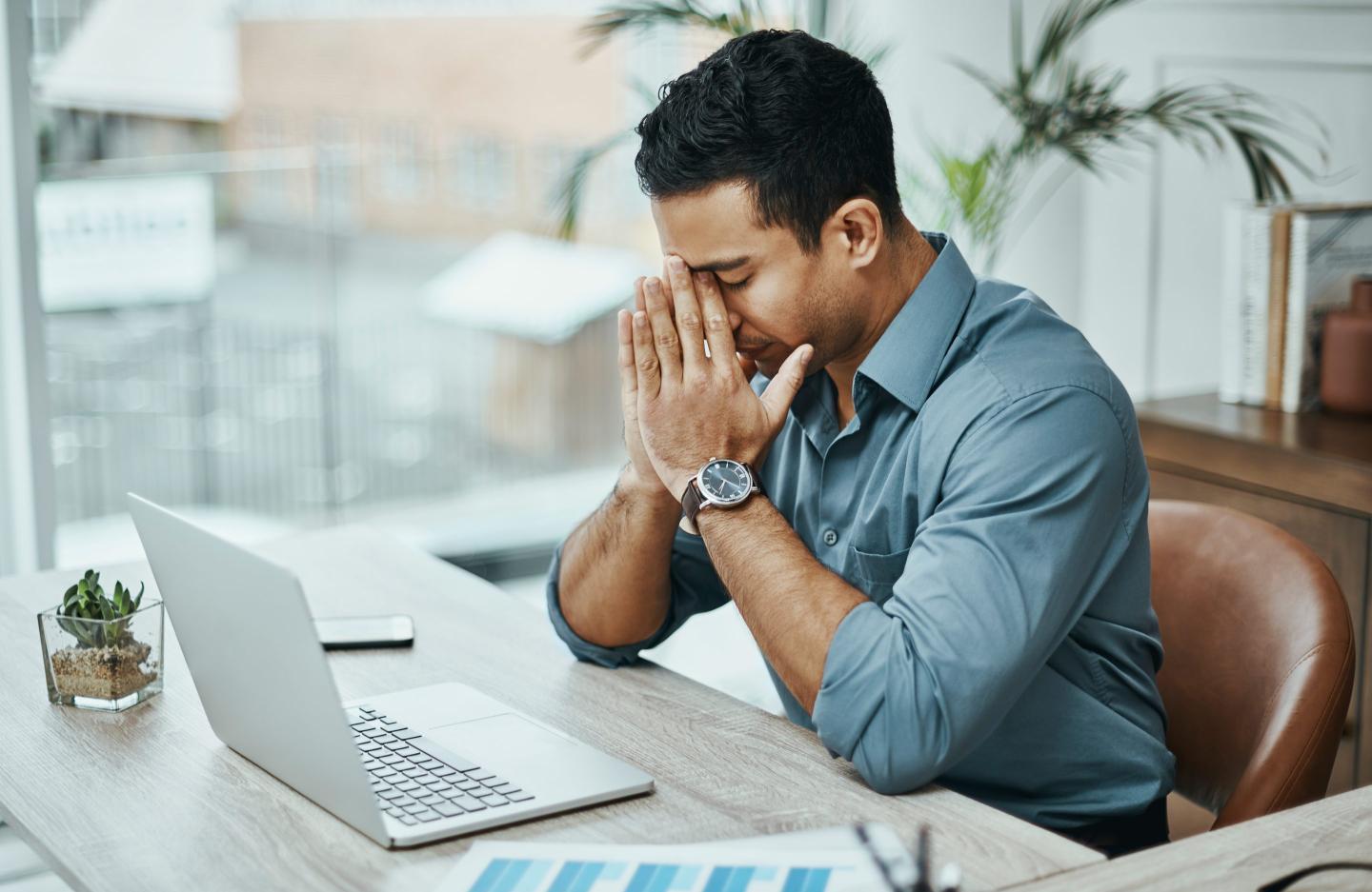 A man sits at a desk with a laptop and has his head in his hands.