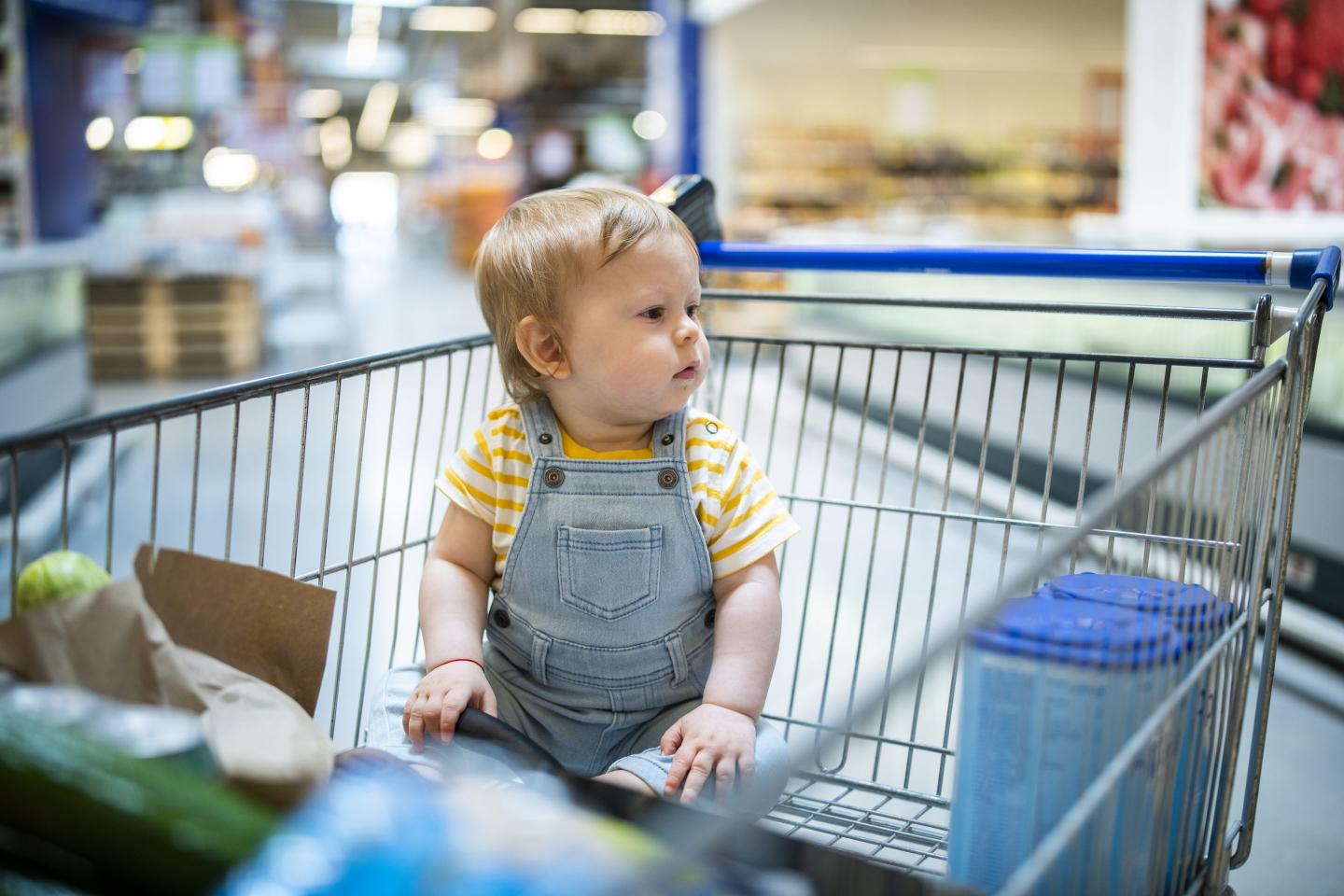 Baby boty sitting in a shopping cart with groceries.