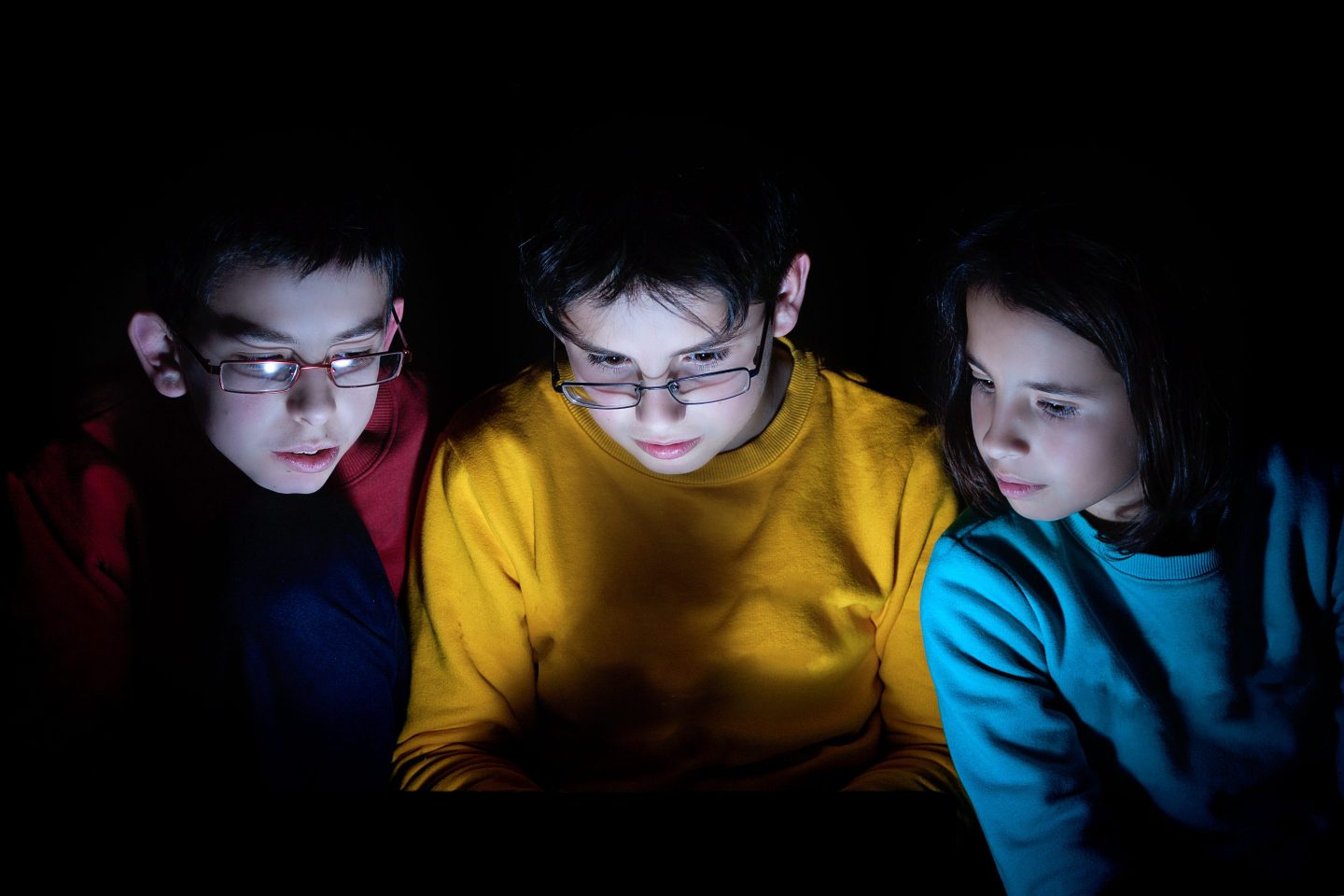 Three brothers are in a completely dark room. Their faces are illuminated by the light of the digital tablet
