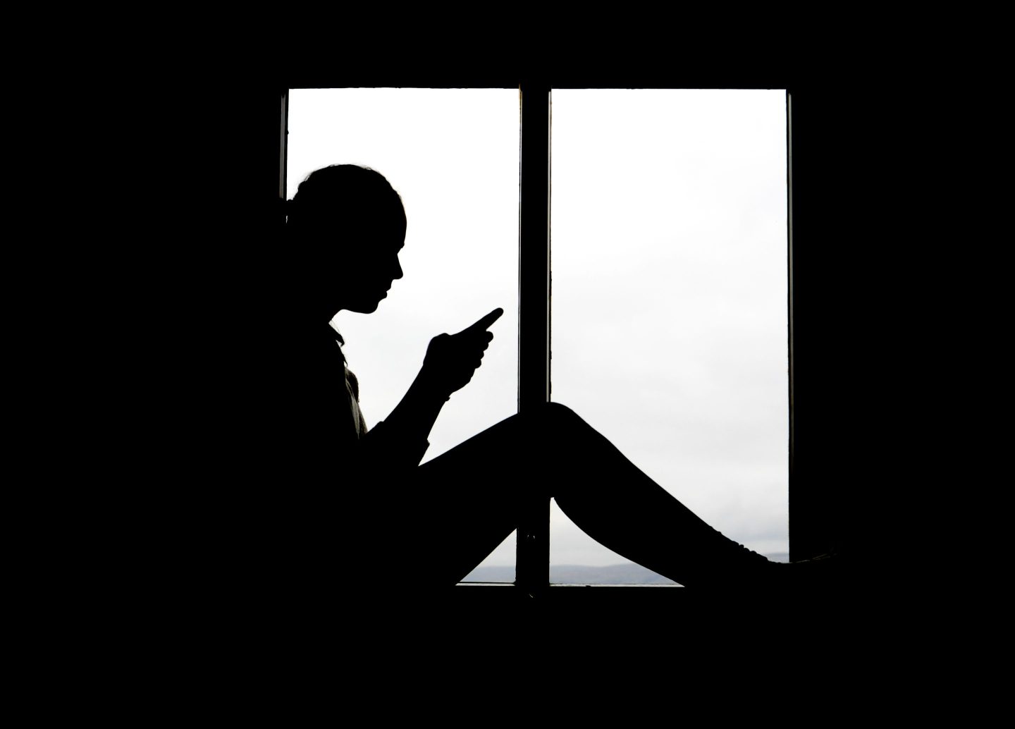 Black and white silhouette of a young girl looking at her phone while sitting in a big window