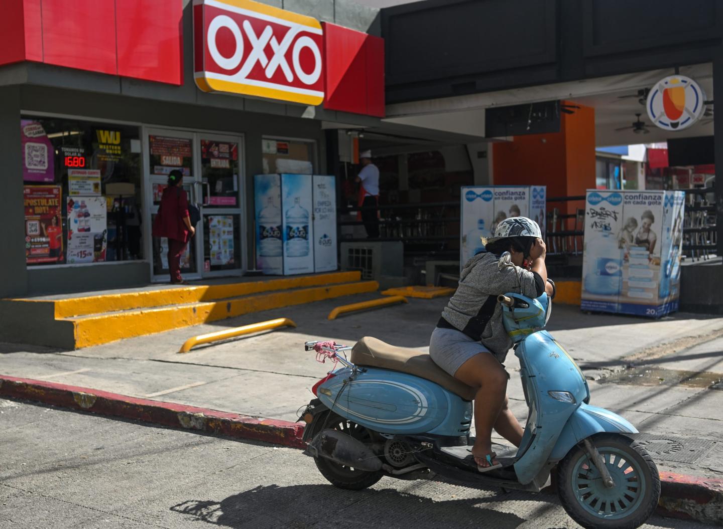 A person sits on a motor scooter outside a gas station in Mexico, looking distressed