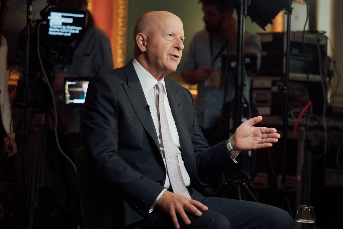 Goldman Sachs CEO David Solomon gestures with his left hand while sitting in front of a digital screen showing illegible text.