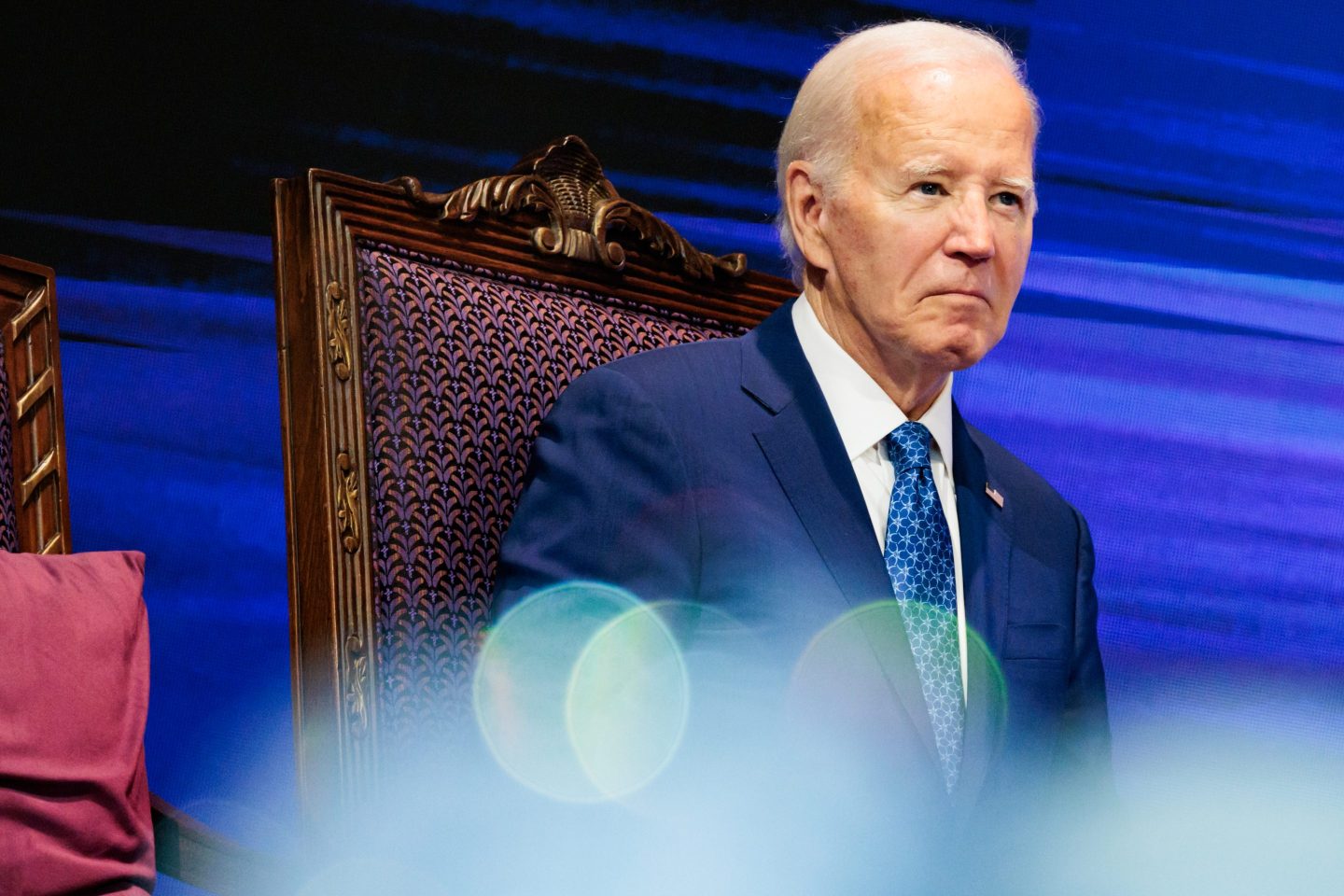 President Biden wearing a navy jacket, white shirt and blue tie sitting in a high-backed chair